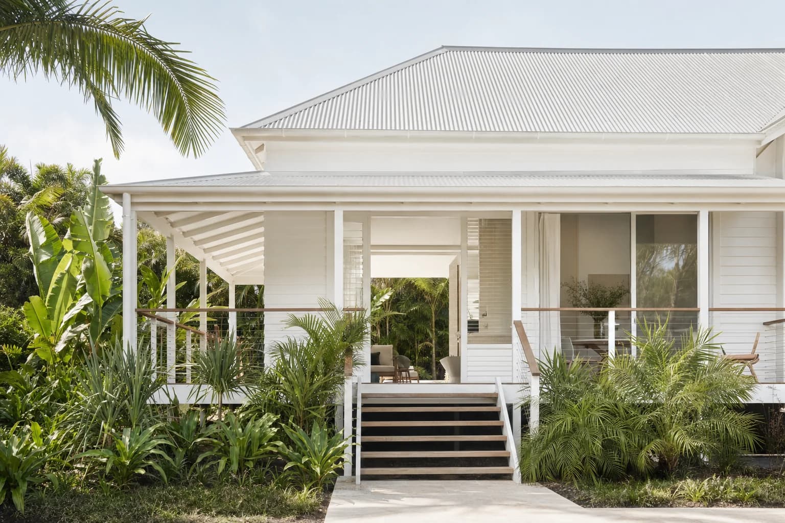 A raised timber beach house on stumps with white weatherboard, a wide front verandah and lush subtropical planting around it