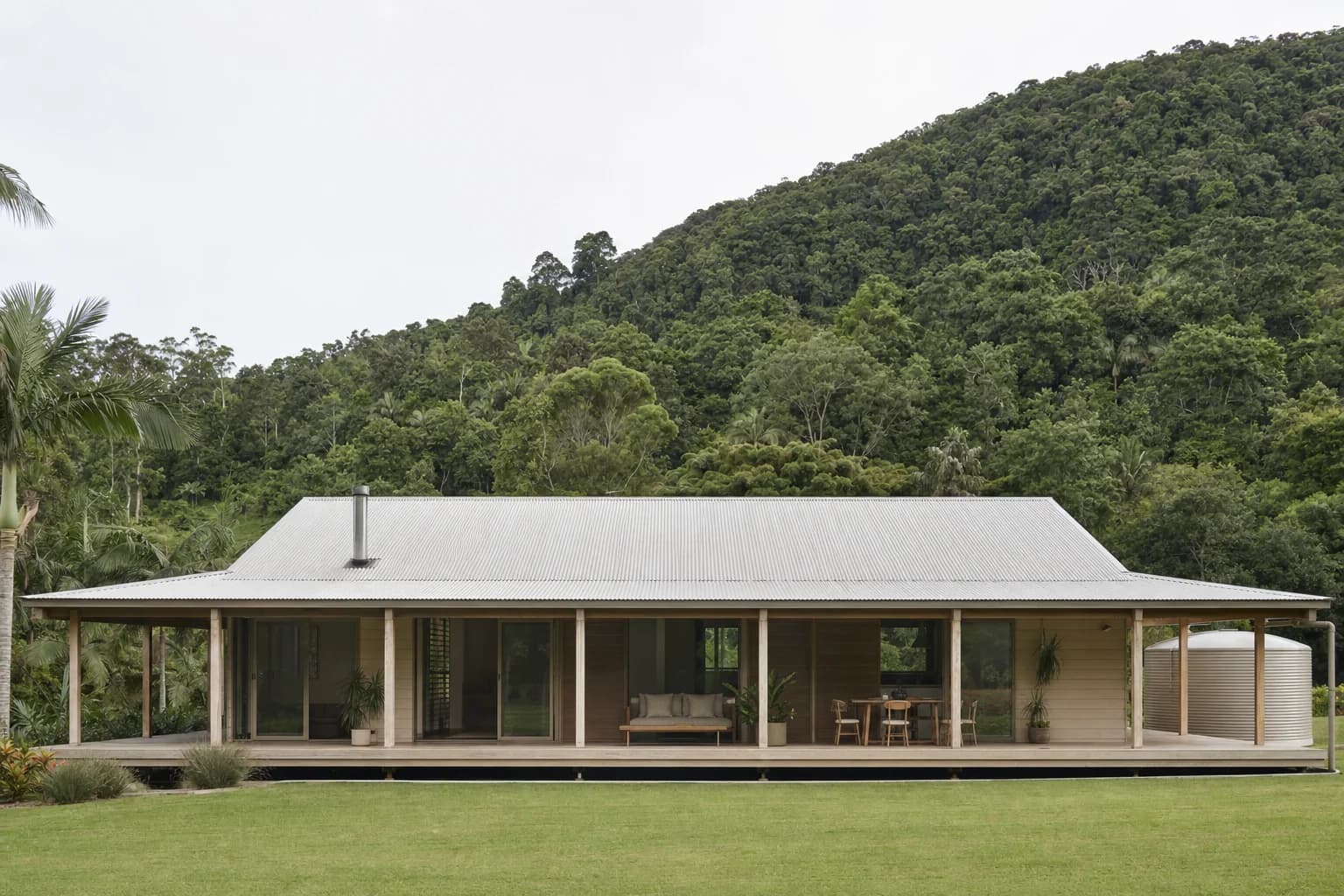 A single storey timber house on a large rural block with a wide verandah, a corrugated metal roof, a rainwater tank and green rainforest hills behind