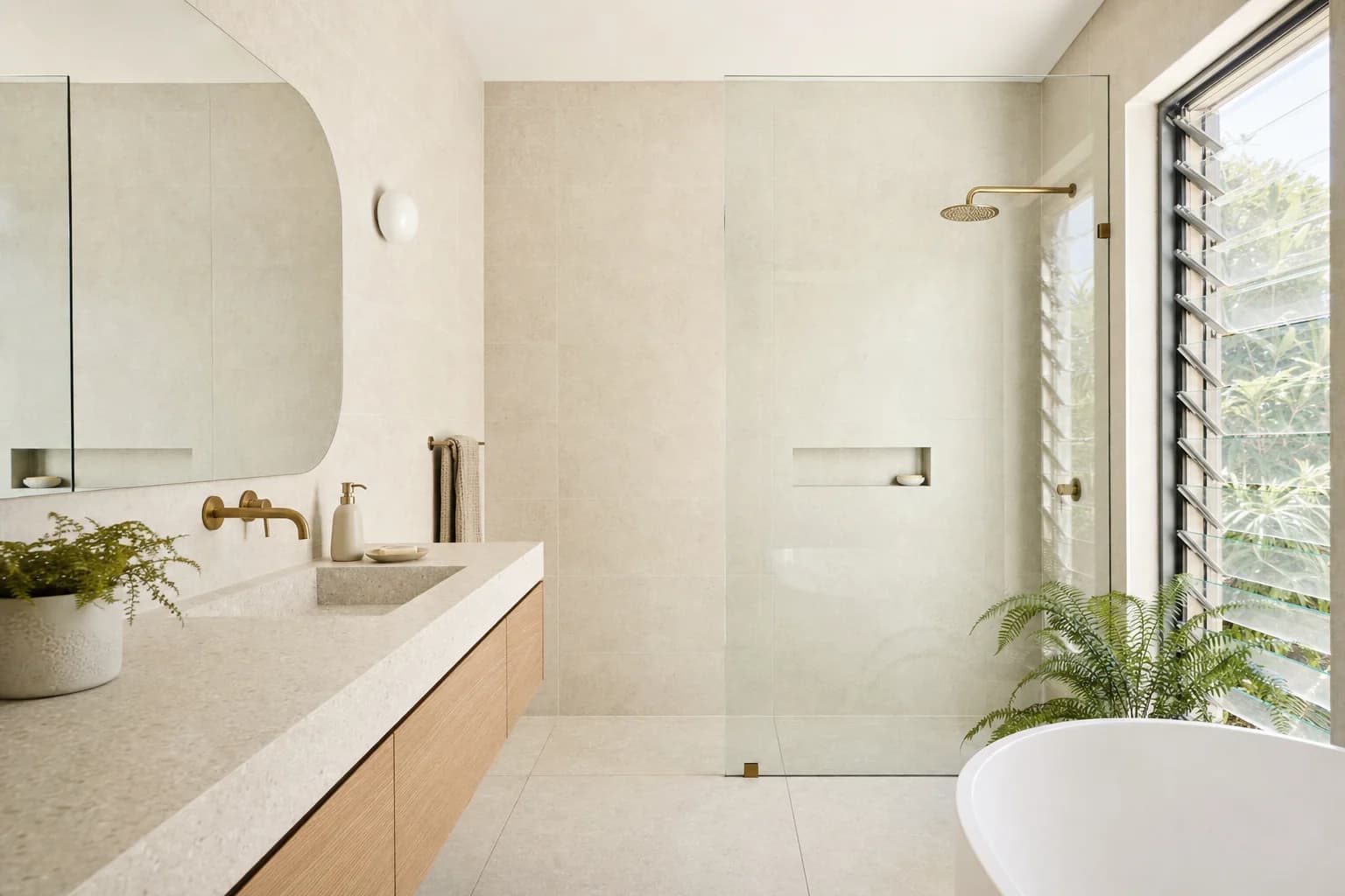 A renovated ensuite in a beach house with warm sand tiling, a slim wall-hung oak vanity, aged brass tapware and a tall louvred window