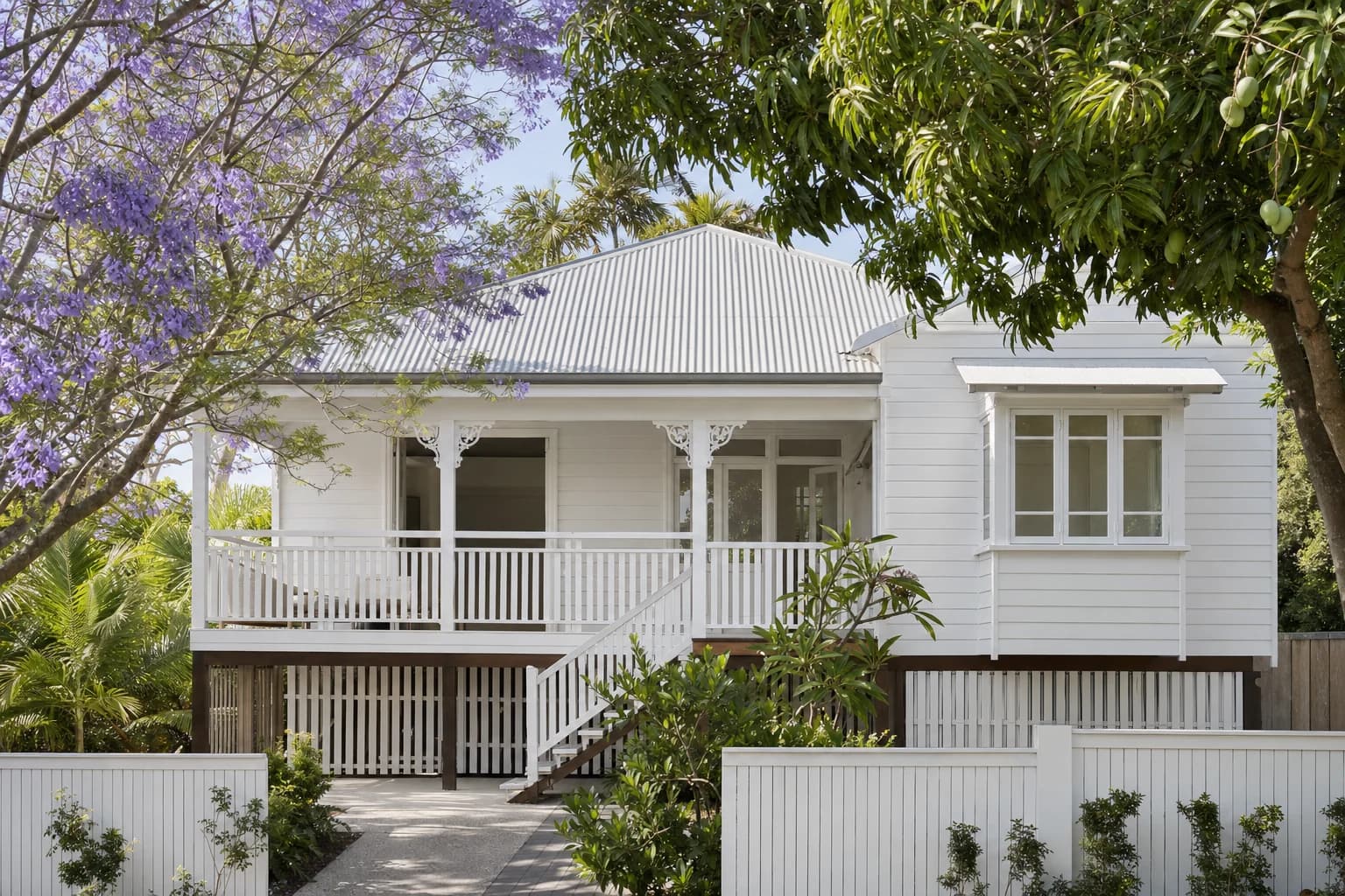 A Queenslander house raised high on timber stumps with white weatherboard, a wide front verandah with decorative brackets and a corrugated metal roof