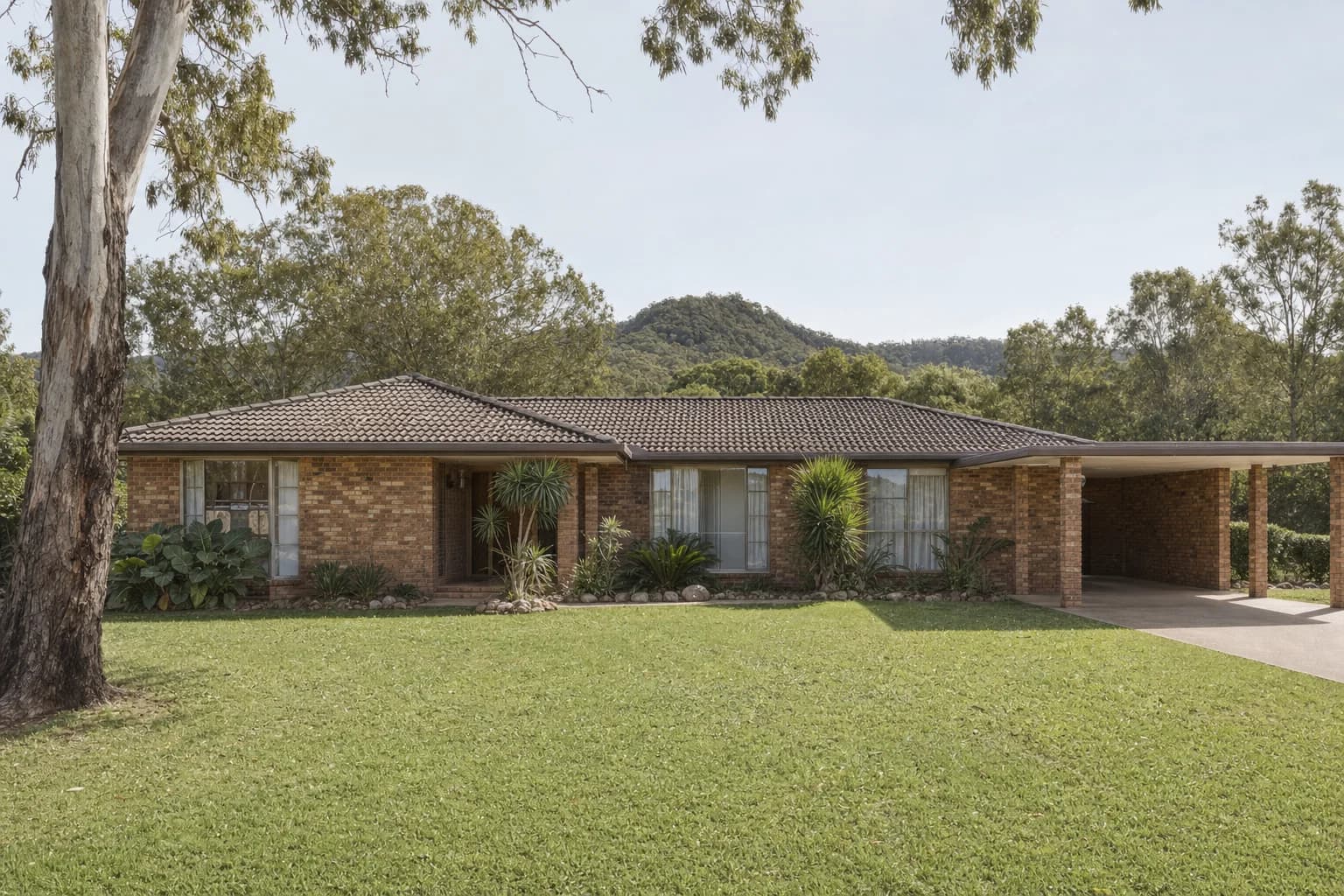 A 1980s low-set face brick house with a tiled hip roof on a large block, a carport to one side and forested hills in the distance